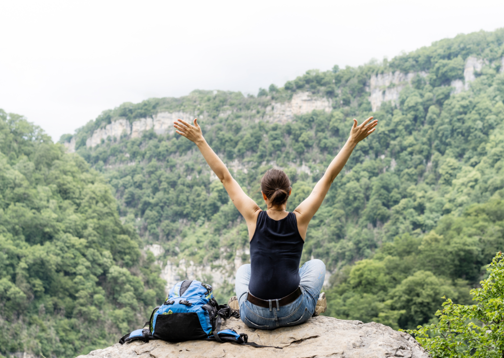 A woman sitting on a cliff with her arms raised, enjoying nature and disconnecting from technology, surrounded by green forested mountains — a moment of “disconnect to reconnect: finding presence beyond the screen.”